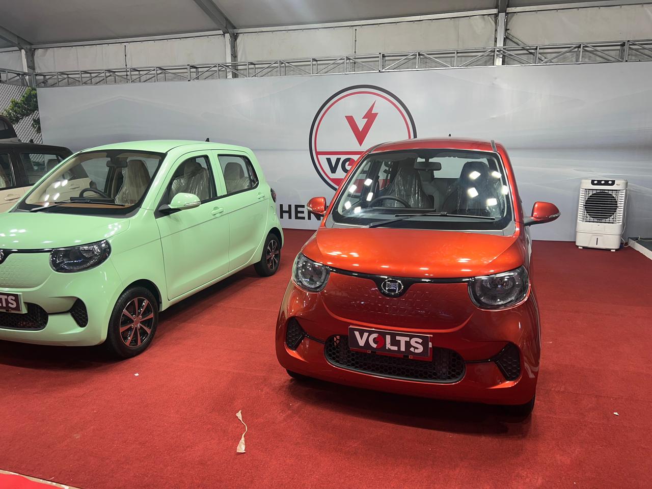 Two VOLTS electric cars, one mint green and one orange, are displayed on a red carpet.