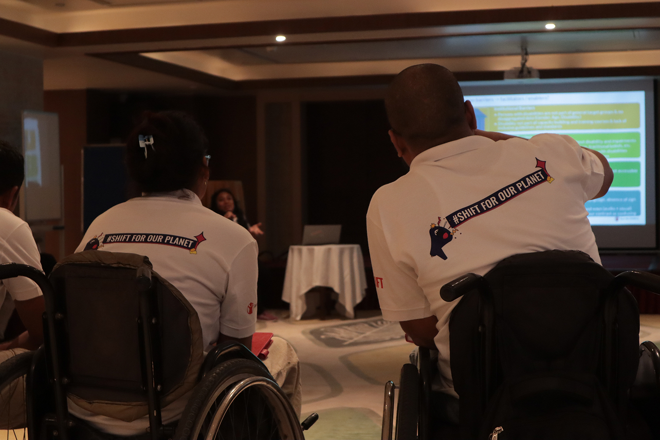 Two individuals in wheelchairs, viewed from behind, attend the "SHIFT FOR OUR PLANET" Youth Advocacy Workshop, looking towards a presenter and a screen displaying information.