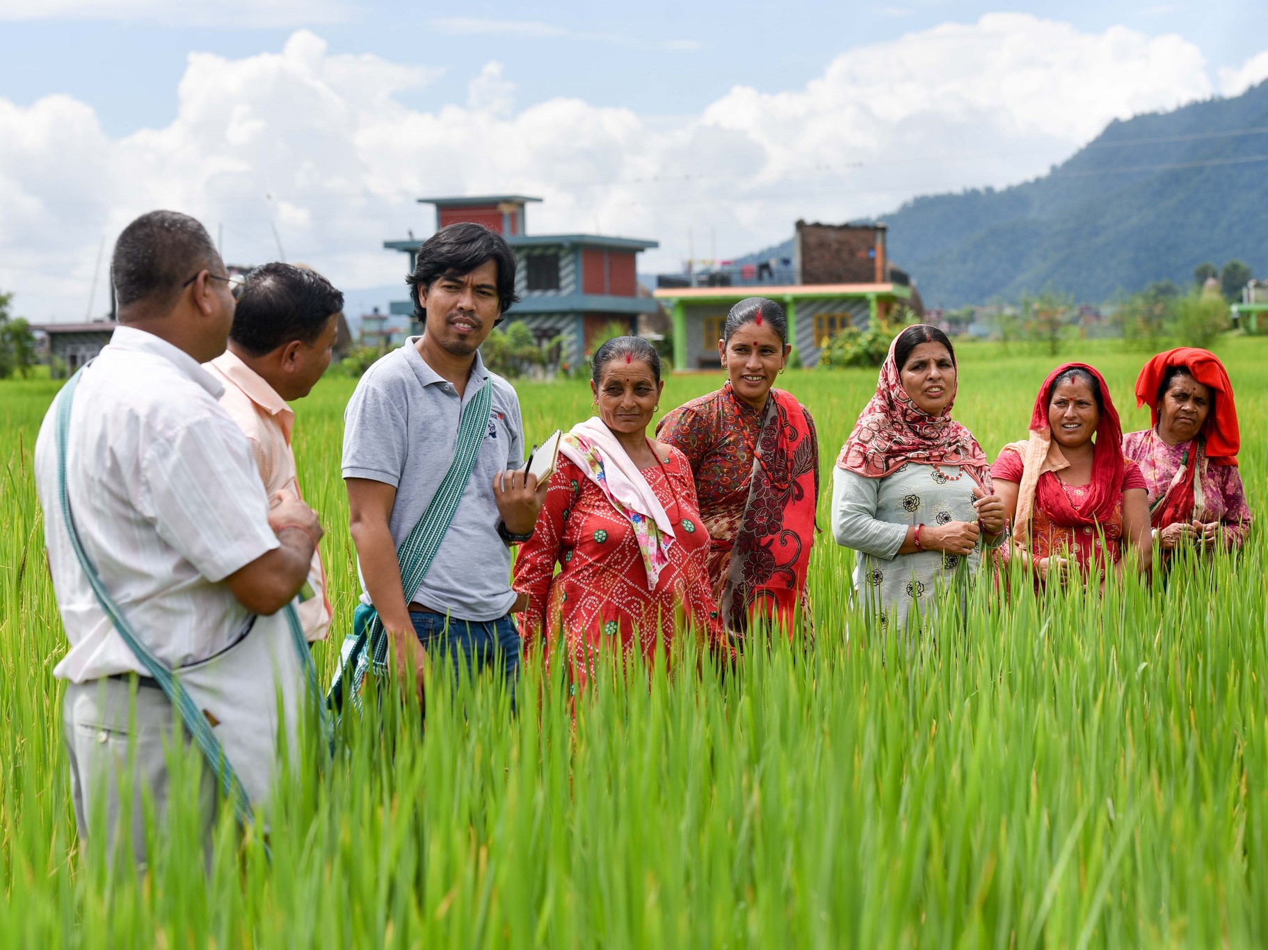 A group of Nepali farmers, including men and women in traditional clothing, are standing in a lush green paddy field, looking toward the camera.