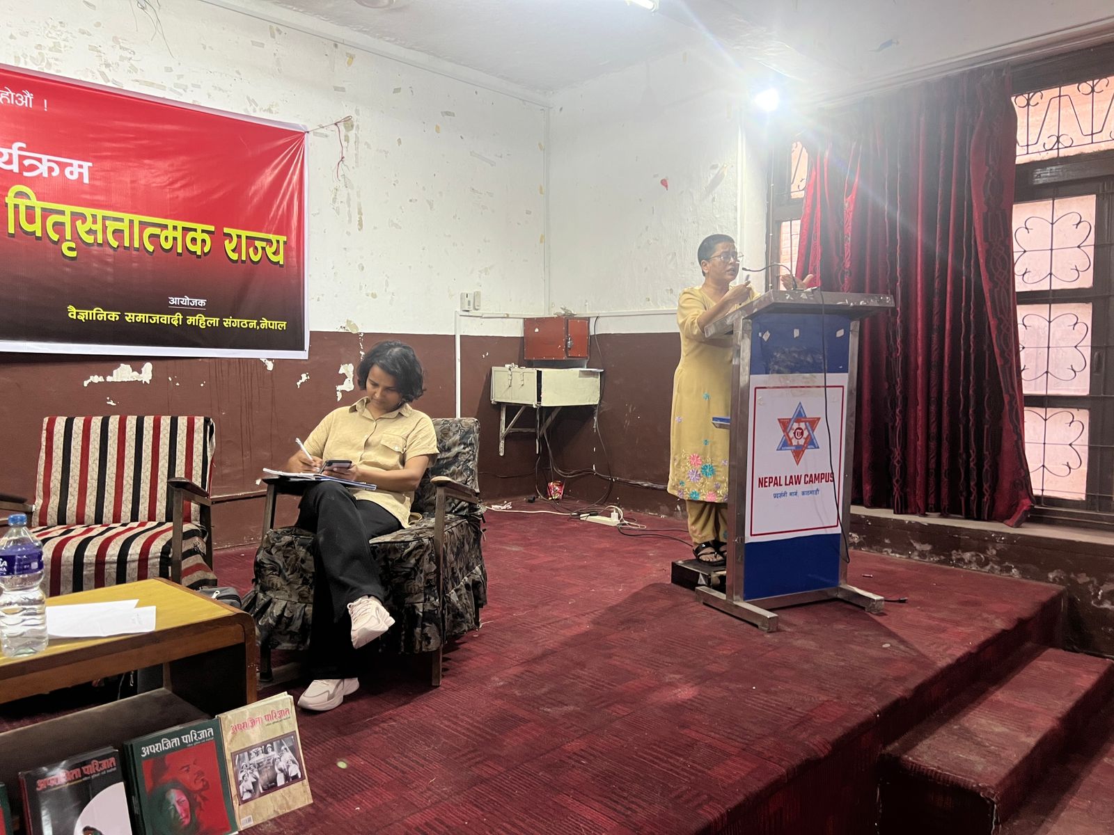 A woman in a yellow outfit is speaking at a podium with the "Nepal Law Campus" logo on it, while a women with short hair sits in a chair taking notes.