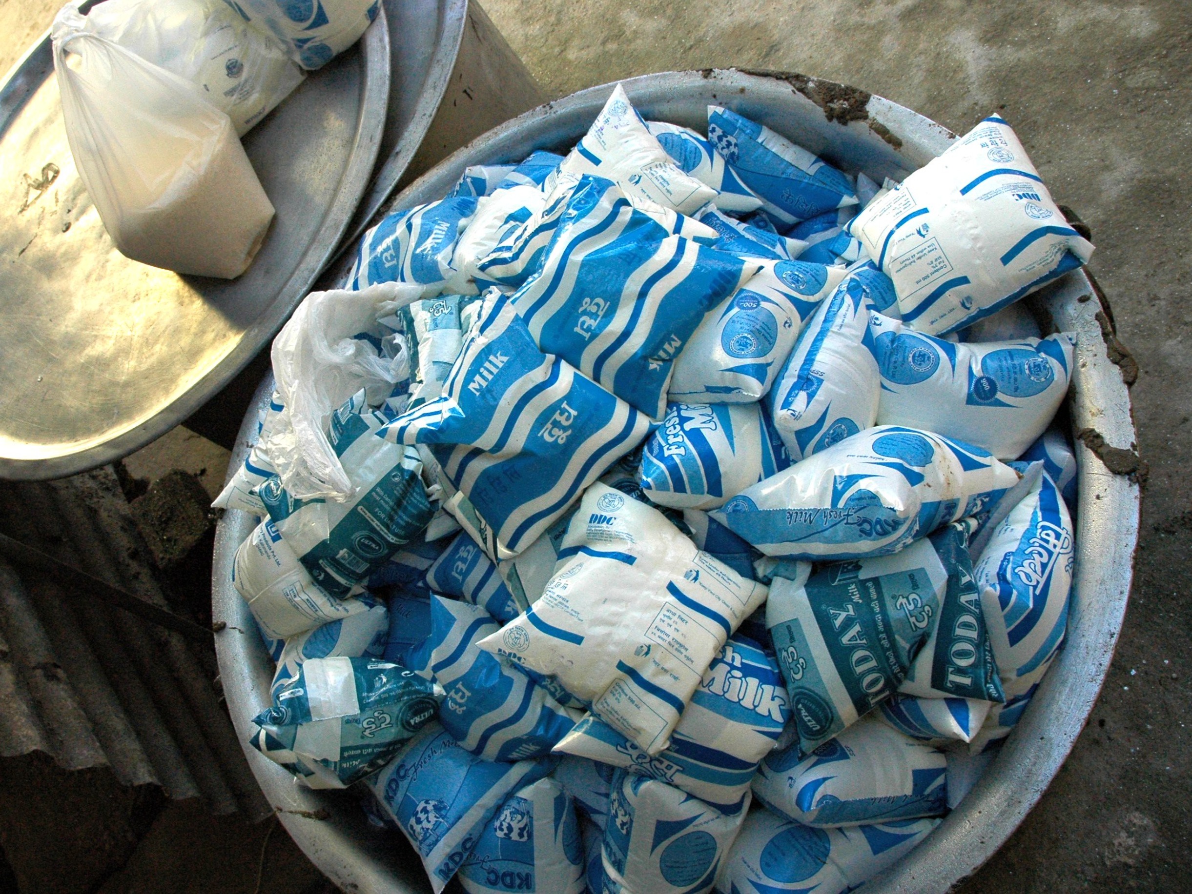 A large aluminum bowl overflowing with plastic bags of milk, with a few bags and a tray visible next to it.
