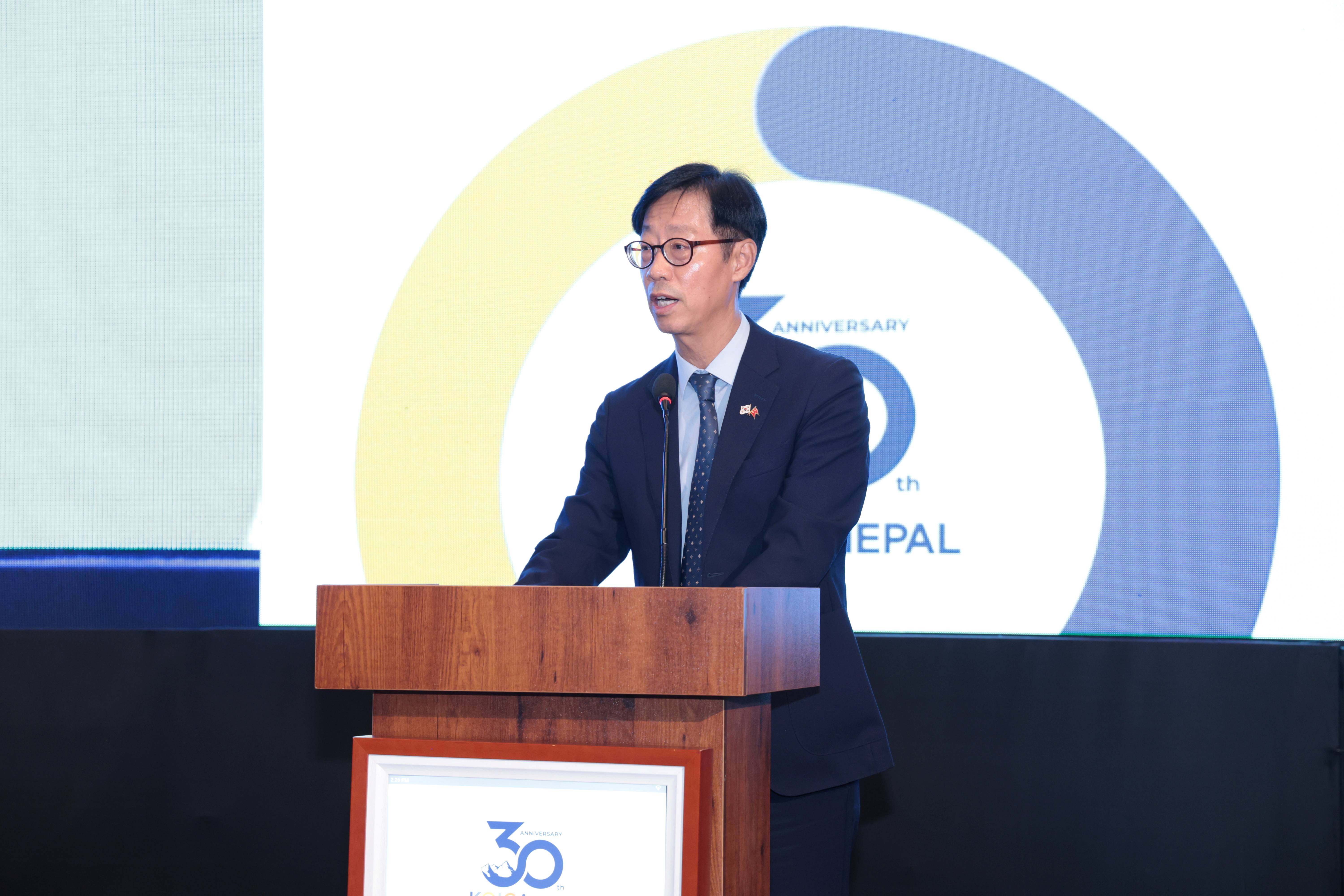 Mooheon Kong, Country Director of KOICA Nepal, delivers his opening remarks at a podium in front of a screen displaying the KOICA Nepal 30th Anniversary logo at Hotel Yak & Yeti, Kathmandu, on August 22, 2025.