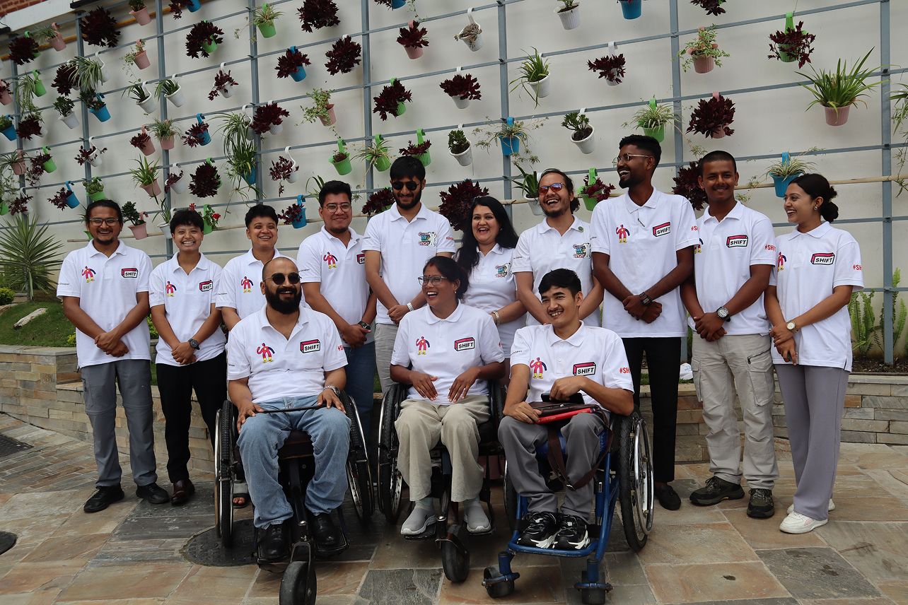 A diverse group of young people, including several in wheelchairs, stand and sit in front of a green wall adorned with potted plants, all smiling and wearing white "SHIFT" branded shirts.