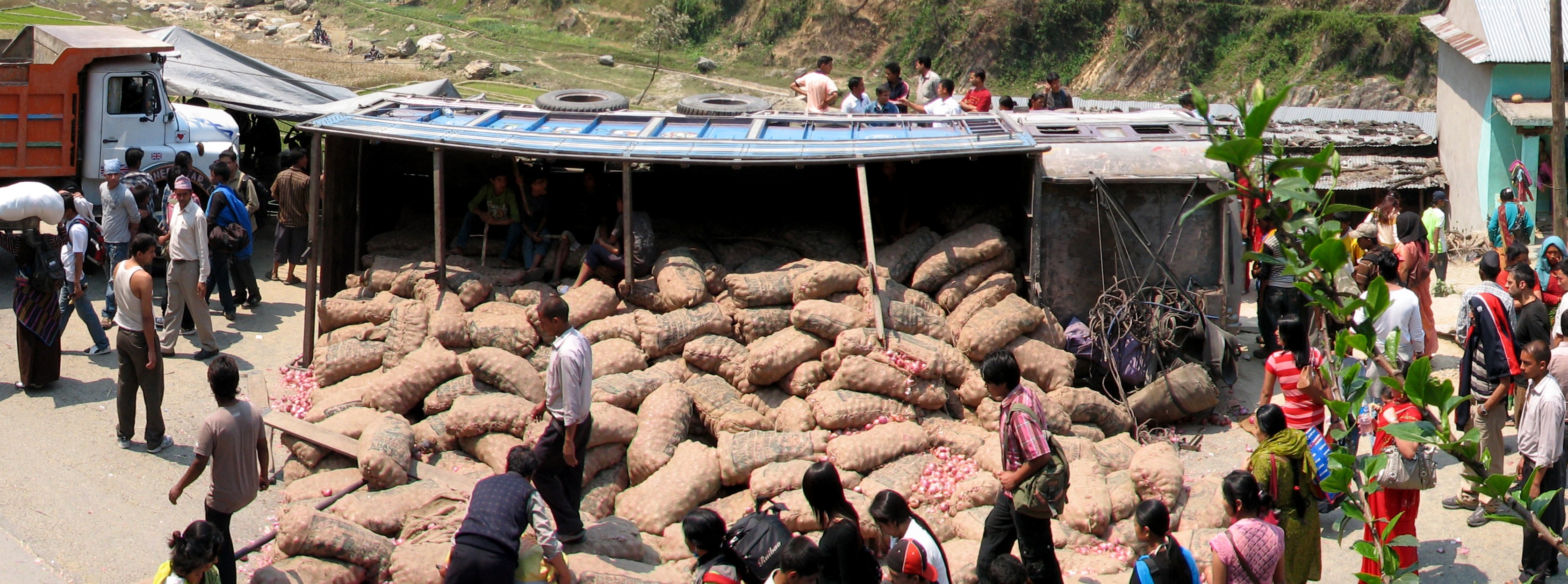 A wide view of a chaotic roadside accident scene with an overturned truck, scattered goods, an orange truck, and numerous onlookers gathered on a dusty road in a rural, hilly area.