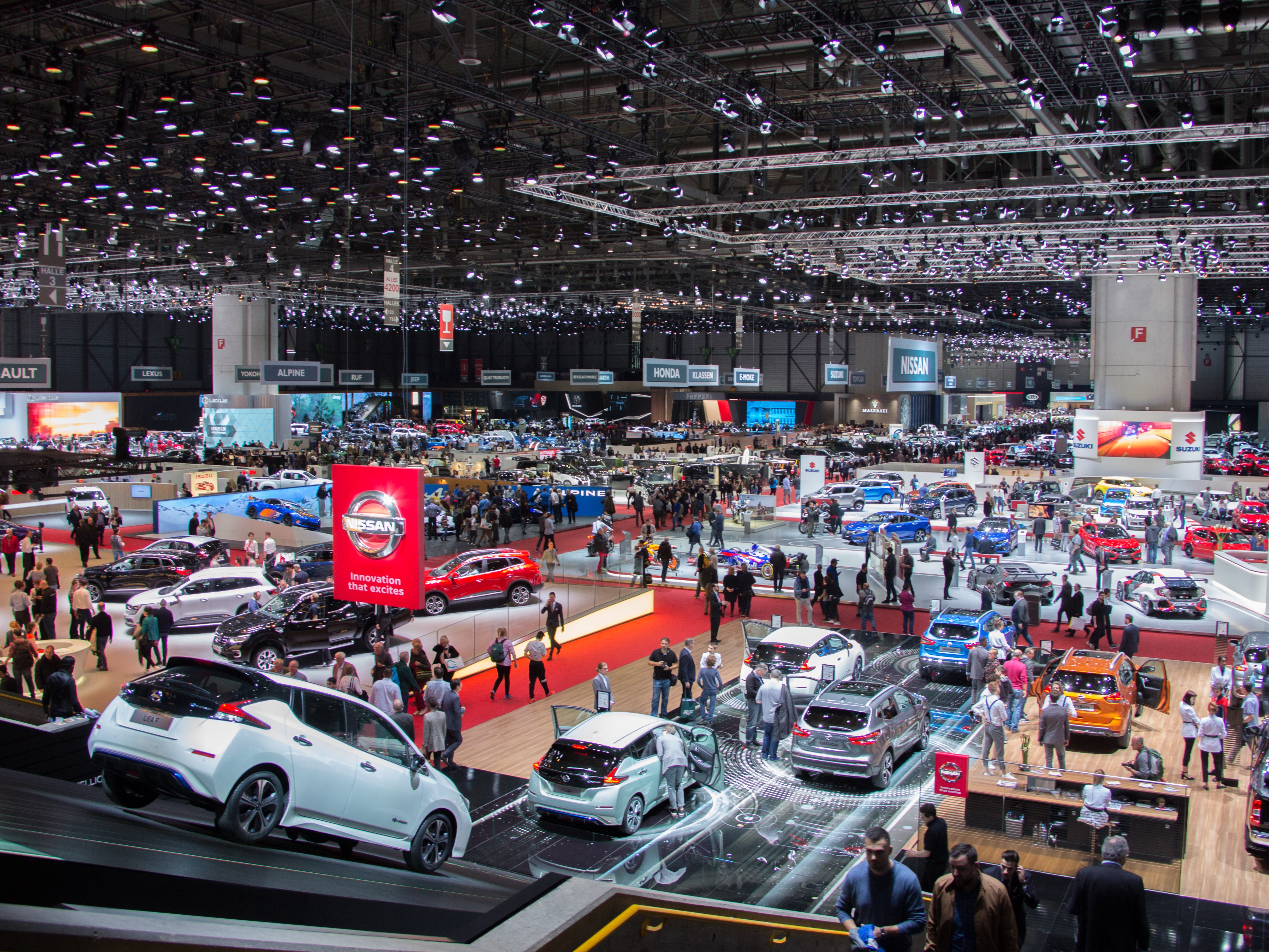 An elevated, wide shot of a massive and bustling auto show. The hall is filled with numerous car displays, including a red Nissan exhibit in the foreground. Crowds of people are visible throughout the space.
