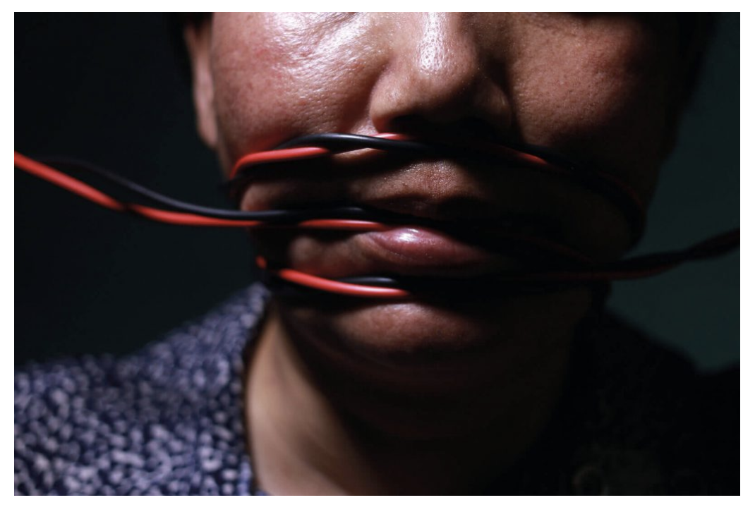 A close-up photograph of a woman's lower face, with black and red electrical wires tightly wrapped across her mouth, symbolizing silence, suppression, and violence.