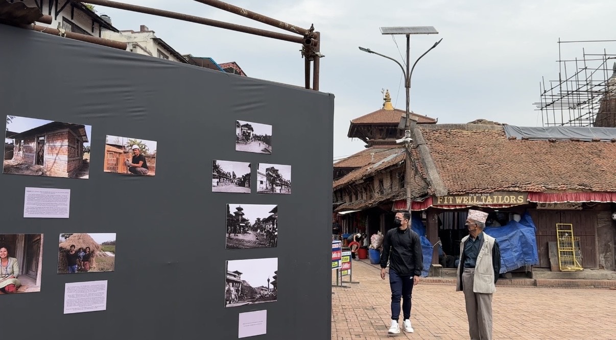 A photo exhibit on a dark gray wall stands in a public square with traditional brick buildings and people walking in the background.