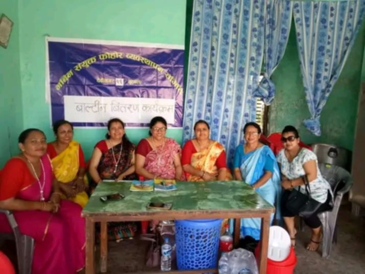 Seven women are seated around a square table during a dustbin distribution event organized by the Nabin Samyukta Waste Management Committee in Devinagar-11, Butwal, with a banner visible in the background.