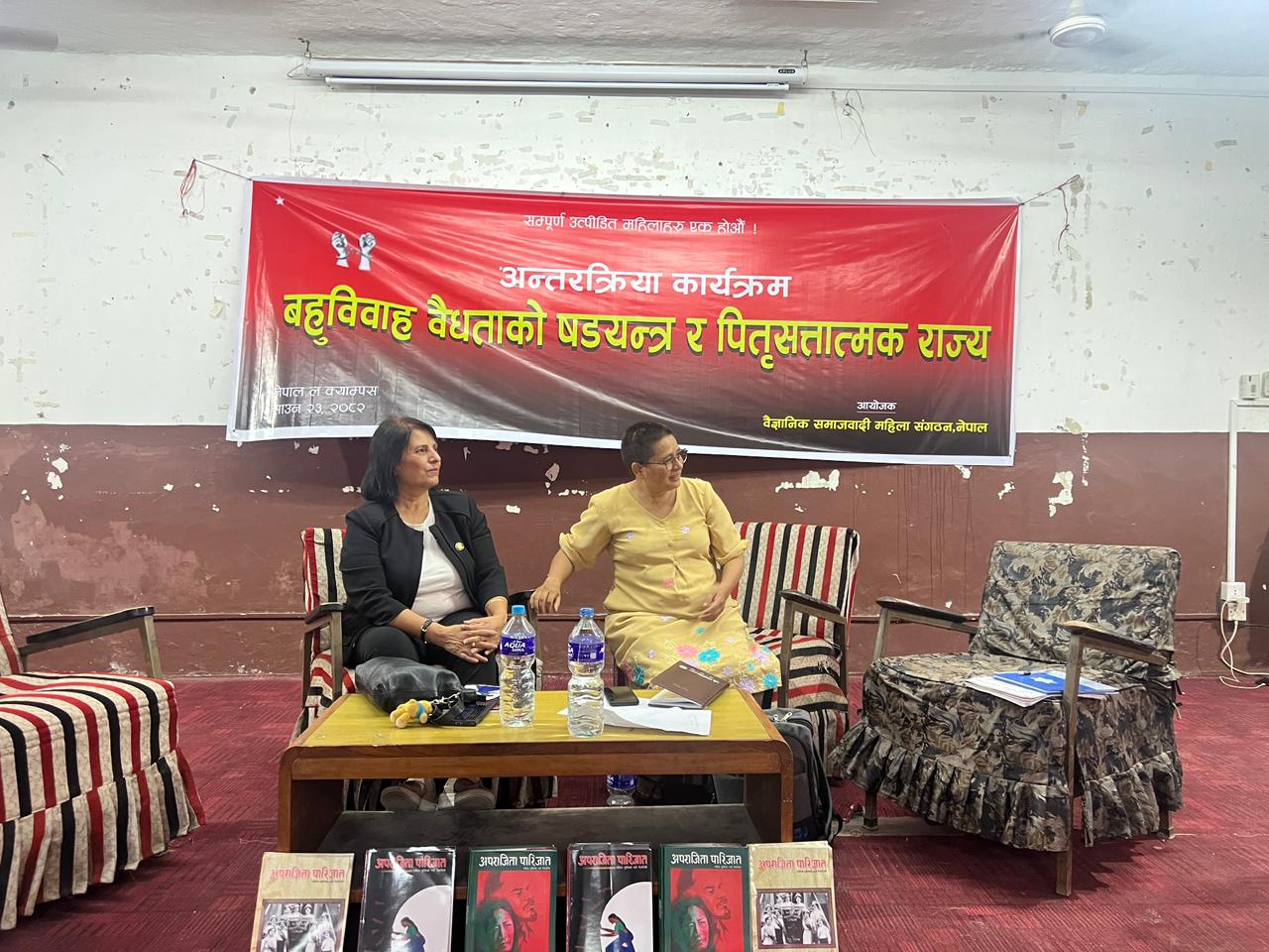 Two women are seated on a platform, with a banner behind them that reads "Interactive Session against Polygamy and Patriarchal State" in Nepali.