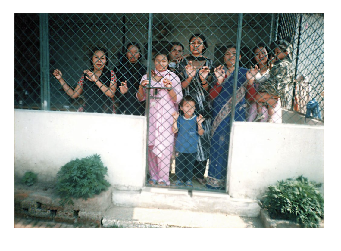 A group of women and two children stand behind a metal gate, looking out from what appears to be a prison or detention facility.
