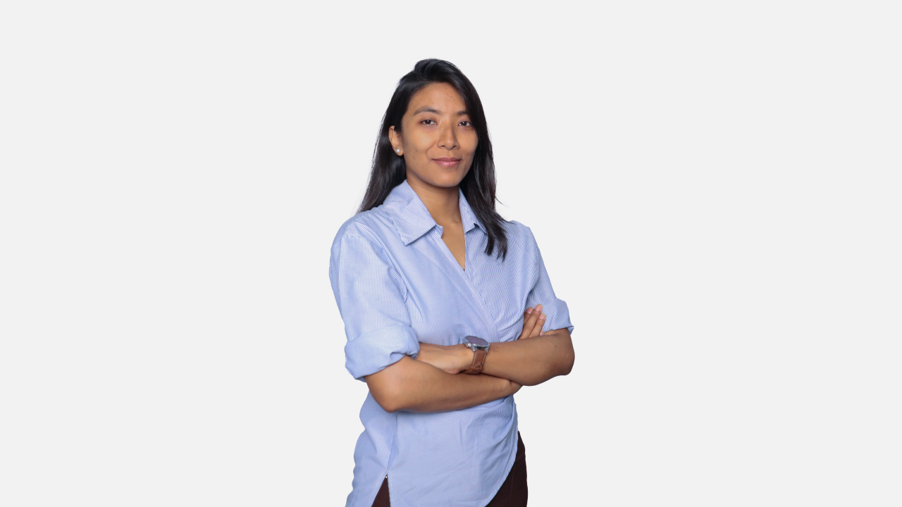 A professional headshot of a young woman with dark hair and a light complexion, wearing a blue collared shirt with her arms crossed, smiling slightly against a plain white background.