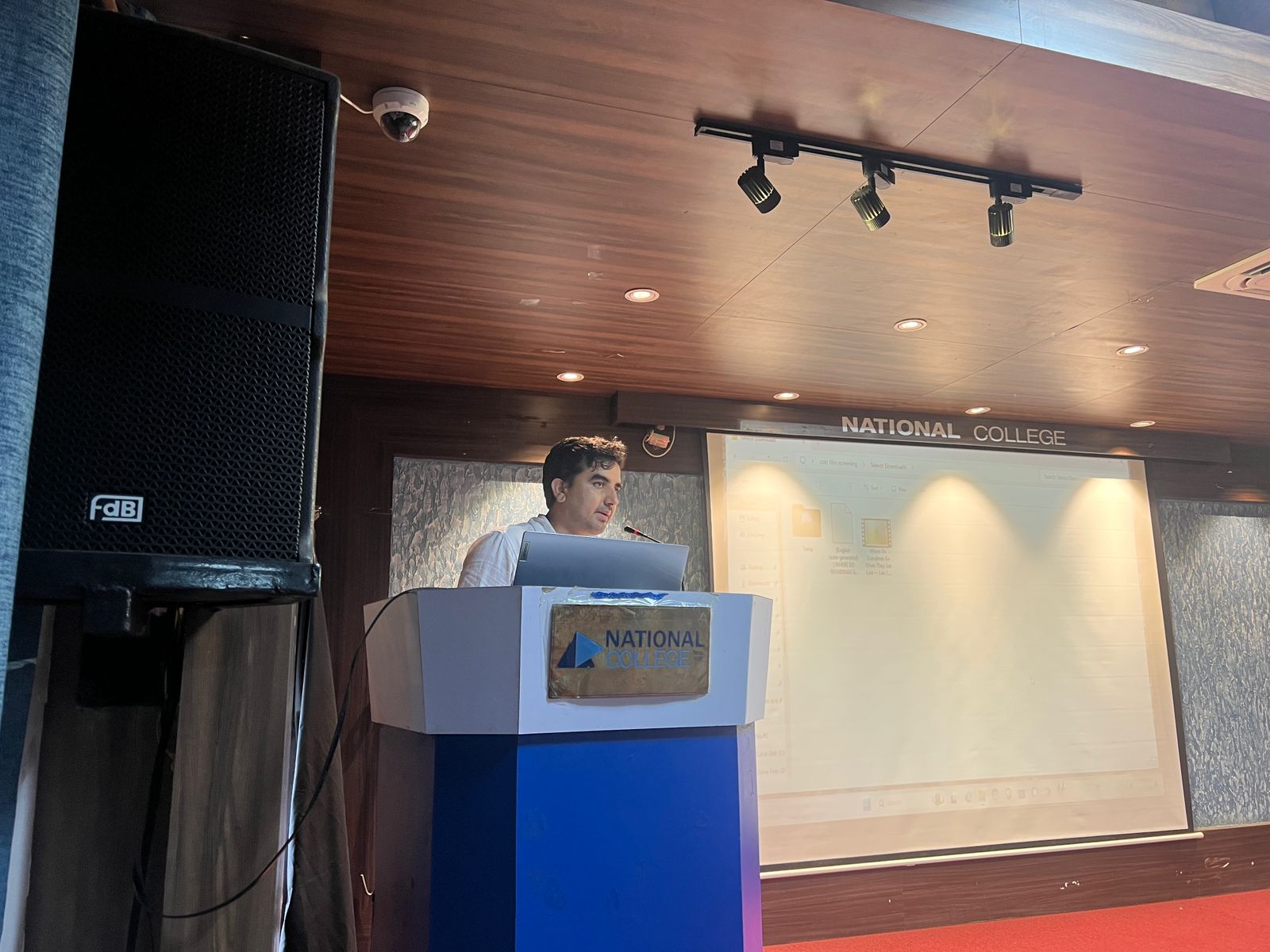 A speaker stands at a podium in front of a projection screen that reads "NATIONAL COLLEGE," addressing an audience at the documentary screening event held at National college on August 4.